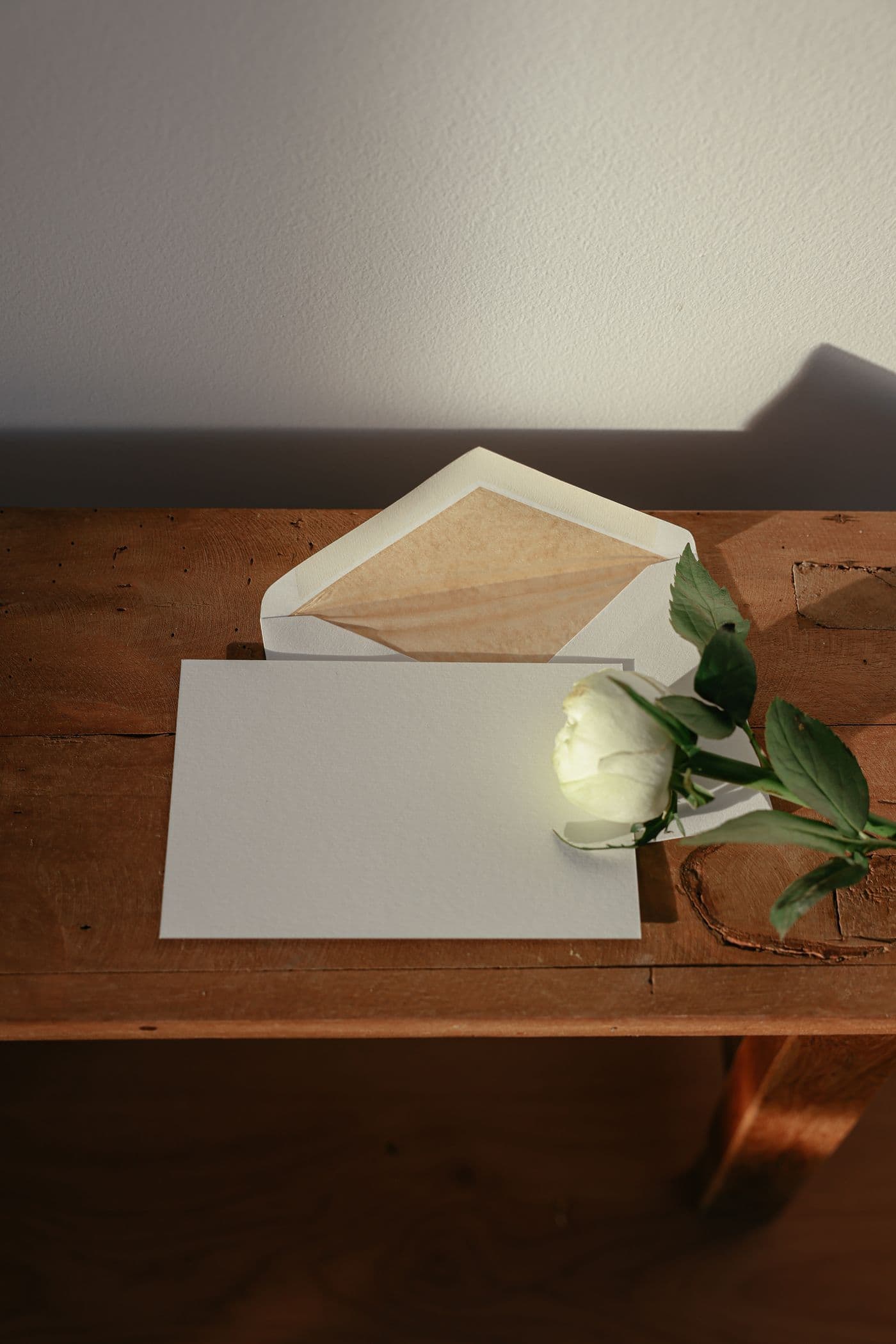 A cream envelope and unwritten card resting on a wooden table beside a single white rose